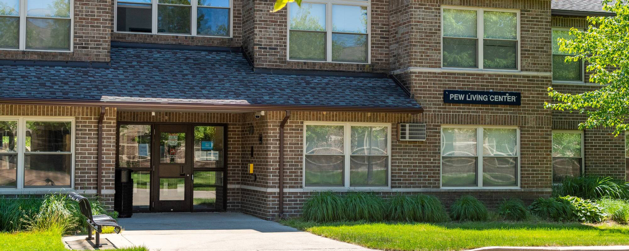 A brick building with a sign reading "Pew Living Center" above the entrance. It has multiple windows and a small path flanked by green bushes and a bench.